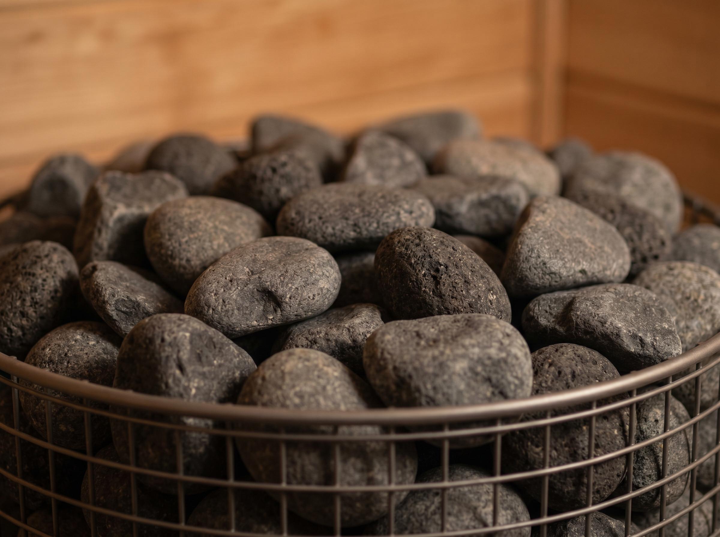 Close-up of a traditional Finnish sauna stove's rock basket filled with dark grey volcanic stones in varied sizes and tones, lit by warm 3000K tungsten light, with Canadian Hemlock timber wall softly defocused in the background.