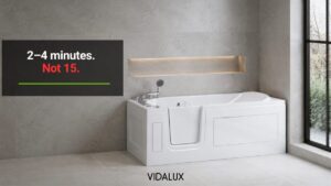 Vidalux Abberton walk-in bath with an inward-opening door installed in a modern grey stone-tiled bathroom, with bare winter trees visible through a large window to the right.